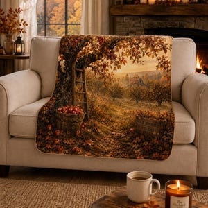 May include: A rectangular throw blanket featuring a printed autumn orchard scene with a large apple tree, a wooden ladder, a basket filled with red apples, and a crate labeled Old Orchard Est 1867, all set against a warm sunset landscape.
