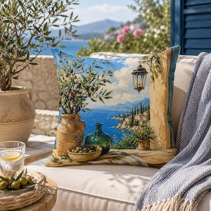 May include: Decorative square throw pillow featuring a scenic coastal landscape with olive branches, a vase, and a green bottle. The pillow has a blue sky and sea background, with a lamp and buildings.