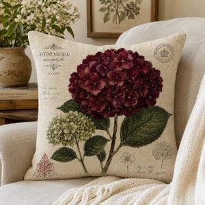 May include: A square throw pillow with a beige linen-look cover featuring a botanical print of a large dark burgundy hydrangea bloom and a smaller green hydrangea cluster, accompanied by vintage-style typography and a circular emblem.
