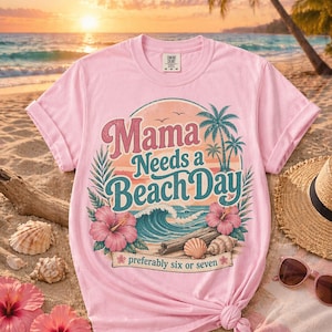 Op de afbeelding: Zachtroze T-shirt met een strandthema. Het ontwerp bevat de tekst "Mama Needs a Beach Day" in roze en blauwgroen, met palmbomen, golven en hibiscusbloemen. De zin "preferably six or seven" is ook opgenomen.