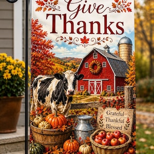 Puede incluir: Una bandera de jardín rectangular con el texto Give Thanks en la parte superior. La escena muestra una vaca blanca y negra, un granero rojo con un silo, calabazas en un barril de madera, una lechera de metal y una cesta de manzanas rojas con un letrero que dice Grateful Thankful Blessed.