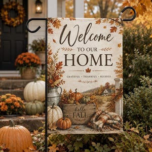 Puede incluir: Una bandera de jardín rectangular con el texto Welcome to our HOME, Grateful, Thankful, Blessed y hello FALL. El diseño muestra una escena rústica con calabazas, una jarra de metal, una caja de madera y una manta a cuadros entre hojas de otoño.