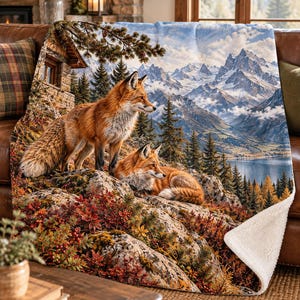 May include: A plush throw blanket featuring a detailed print of two red foxes resting on a rocky, foliage-covered hillside. The background displays a rustic wooden cabin, a serene lake, and snow-capped mountain peaks under a bright sky.