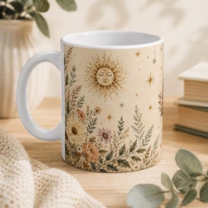 May include: A white ceramic mug featuring a beige background with a printed illustration of a sun with a human face, surrounded by various wildflowers, ferns, and small star shapes scattered across the surface of the mug.