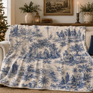May include: A cream-colored throw blanket featuring a blue toile-style print depicting a nativity scene with figures, camels, palm trees, and angels. The detailed pattern shows a central stable, shepherds, and a star in a landscape setting.