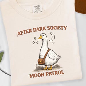 Pode incluir: Uma t-shirt de cor creme com um gráfico de um ganso branco a usar uma mala castanha, acompanhado por uma lua crescente e estrelas. O texto AFTER DARK SOCIETY está acima do ganso e MOON PATROL abaixo em letras de cor castanha.