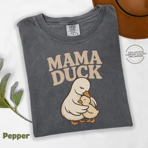 May include: A dark grey t-shirt featuring the text "MAMA DUCK" in tan, with an illustration of a mother duck and duckling. The shirt is labelled "Pepper" and made of pre-shrunk, 100% cotton Comfort Colors.