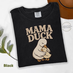 May include: Black t-shirt featuring the text "MAMA DUCK" in tan, above a graphic of a mother duck and duckling. The shirt is made of 100% cotton and is pre-shrunk.