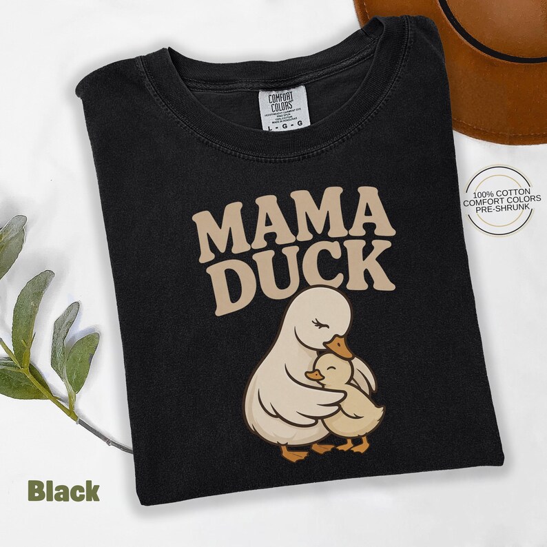 May include: Black t-shirt featuring the text "MAMA DUCK" in tan, above a graphic of a mother duck and duckling. The shirt is made of 100% cotton and is pre-shrunk.