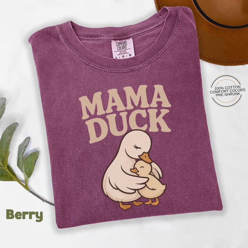 May include: Berry-coloured t-shirt featuring the text "MAMA DUCK" and an illustration of a mother duck and duckling. The shirt is made of 100% cotton and pre-shrunk. A brown hat is in the background.