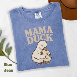 May include: Blue jean-coloured t-shirt with the words "MAMA DUCK" in tan lettering. The shirt features an illustration of a mother duck embracing a baby duck. The shirt is a Comfort Colors brand, size L-G-G.