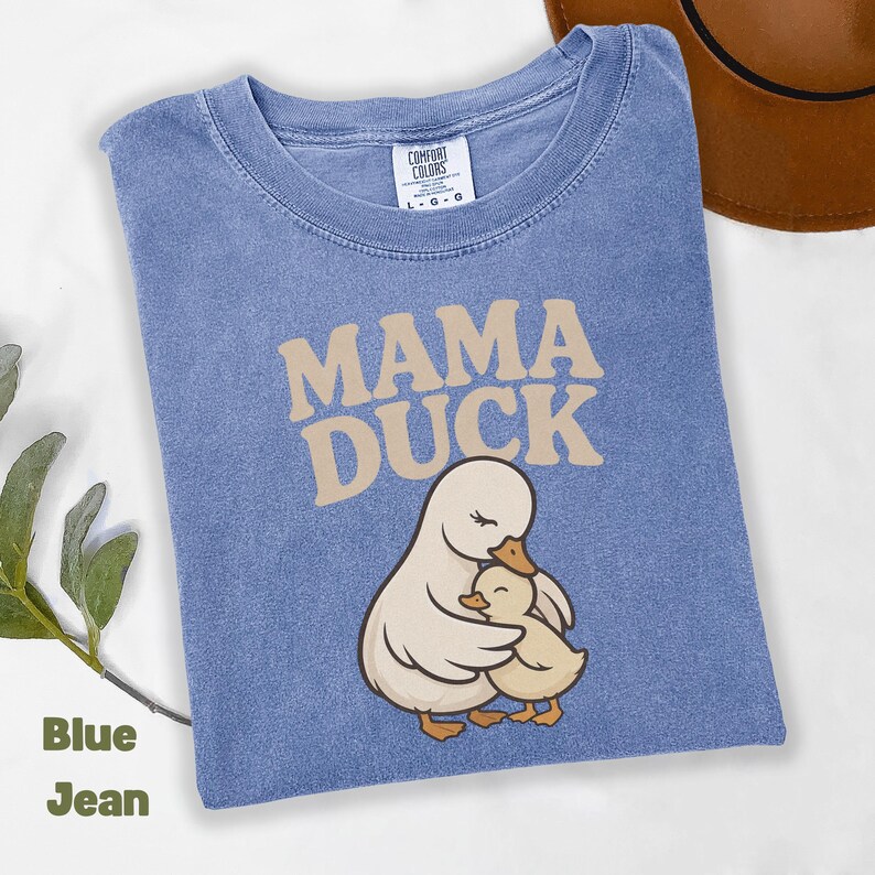 May include: Blue jean-coloured t-shirt with the words "MAMA DUCK" in tan lettering. The shirt features an illustration of a mother duck embracing a baby duck. The shirt is a Comfort Colors brand, size L-G-G.