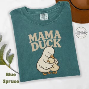 May include: Blue Spruce t-shirt featuring the text "MAMA DUCK" and an illustration of a mother duck and duckling. The shirt is made of 100% cotton and is pre-shrunk. The t-shirt is a Comfort Colors brand.