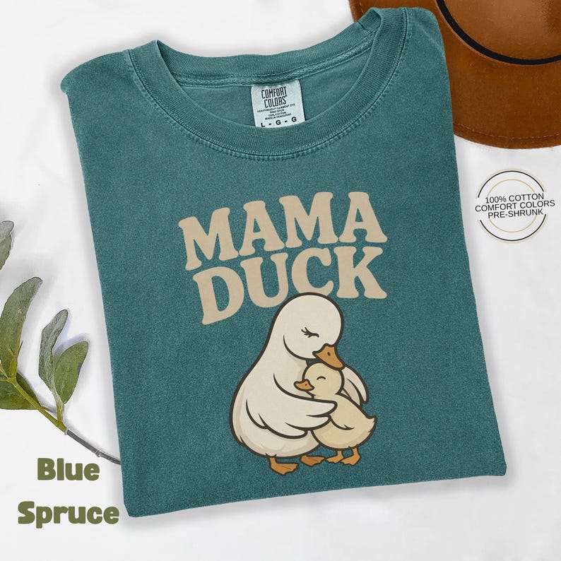 May include: Blue Spruce t-shirt featuring the text "MAMA DUCK" and an illustration of a mother duck and duckling. The shirt is made of 100% cotton and is pre-shrunk. The t-shirt is a Comfort Colors brand.