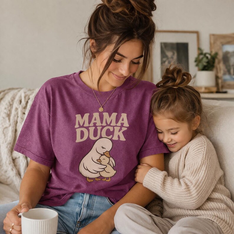 May include: A plum-coloured t-shirt featuring the text "MAMA DUCK" in a beige font, and a graphic of a mother duck and duckling. The shirt is worn by a person, with a child beside them.