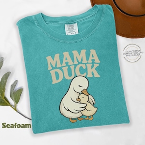 May include: Seafoam green t-shirt with the words "MAMA DUCK" in beige lettering. The shirt features an illustration of a mother duck embracing a baby duck. The shirt is made of 100% cotton and is pre-shrunk.