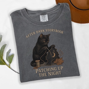 Könnte beinhalten: Ein anthrazitfarbenes Kurzarm-T-Shirt mit einer Grafik einer schwarzen Katze, die einen dunklen Stoffflicken mit Mondsichel und Sternen näht. Der Text lautet AFTER DARK STORYBOOK und PATCHING UP THE NIGHT. Das Shirt hat ein Comfort Colors Etikett.
