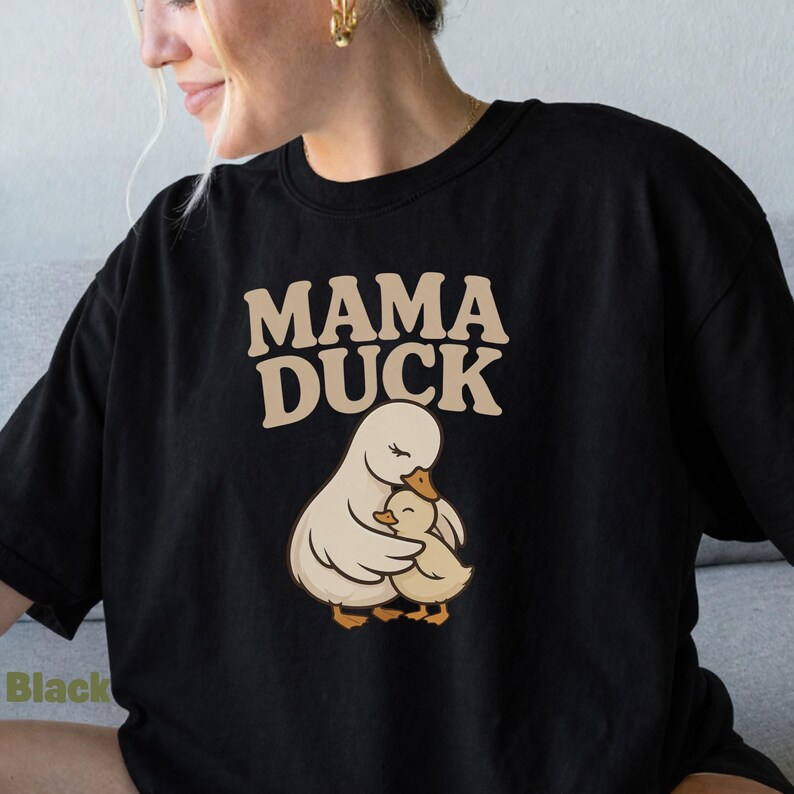 May include: Black t-shirt featuring the text "MAMA DUCK" in a beige, rounded font. Below the text is an illustration of a mother duck embracing a baby duck. The ducks are beige and yellow.
