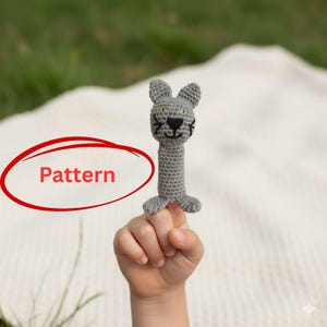Könnte beinhalten: Ein graues gehäkeltes Katzenspielzeug mit schwarzen, gestickten Gesichtszügen und kleinen Ohren. Das Spielzeug wird von der Hand eines Kindes gehalten. Das Wort "Pattern" ist rot umkreist.