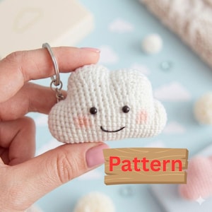 Könnte beinhalten: Ein weißer Häkel-Wolken-Schlüsselanhänger mit einem lächelnden Gesicht und rosigen Wangen. Der Schlüsselanhänger hat einen silbernen Ring und wird in einer Hand gehalten. Das Wort "Pattern" befindet sich auf einem Holzschild im Bild.