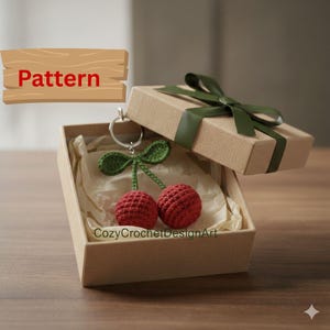 Puede incluir: Un llavero de cerezas de ganchillo en una caja de regalo. El llavero presenta dos cerezas rojas con hojas verdes y un anillo plateado. La caja de regalo marrón está atada con una cinta verde. La palabra "Pattern" es visible.