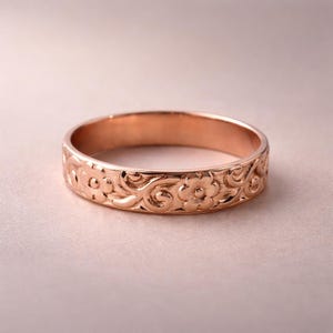 Könnte beinhalten: Ein roségoldener Ring mit floralem Muster. Der Ring hat ein sich wiederholendes Design aus Blumen und wirbelnden Blättern. Das Band ist etwa 0,5 cm breit und hat eine polierte Oberfläche. Der Ring ist vor einem weichen rosa Hintergrund platziert.