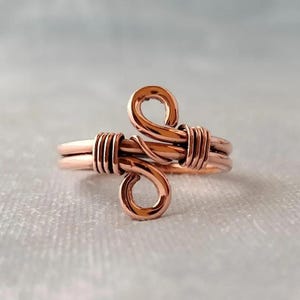 May include: A handcrafted copper wire ring with a unique design. The ring features two looped elements and wrapped wire detailing. The copper has a warm, reddish-brown hue, and the ring is set against a soft, neutral background.