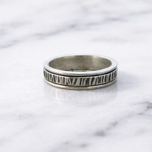 May include: A silver ring with a textured band. The ring has a simple, classic design, with a band that features a series of vertical lines. The ring is likely made of sterling silver or a similar metal.