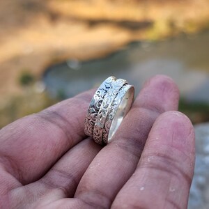 May include: A silver ring with an intricate floral pattern. The ring has a spinning band, and is held in a hand. The ring's design features detailed carvings, and the metal has a polished finish. The ring is a piece of jewelry.