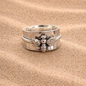 May include: A silver-colored ring with a detailed bee design. The ring features a wide band with a spinning outer ring, and the bee is centered on the front. The ring is set on a sandy surface.
