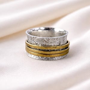 May include: A silver and gold-tone spinner ring. The ring features a textured silver band with an etched design and three gold-tone bands. The ring is set against a white, silky background.