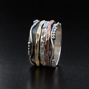 May include: A multi-band ring with a silver base and several textured bands in gold, silver, and copper tones. The ring has a unique design with a mix of smooth and patterned surfaces, creating a visually appealing piece of jewelry.
