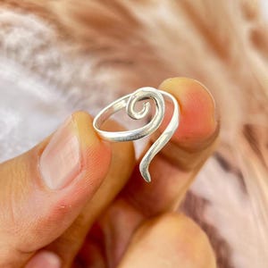 Puede incluir: Un anillo de tono plateado que presenta una banda continua que se curva en un diseño de espiral prominente en la parte superior. El metal tiene un acabado liso y pulido, y la estructura abierta permite que la espiral sobresalga un poco.