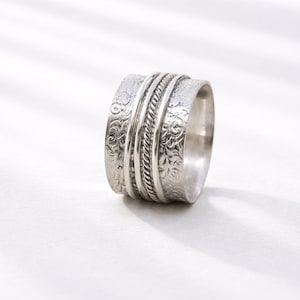 May include: A silver-colored ring with an intricate design. The ring features a series of textured bands, including floral patterns and a braided rope-like detail. The ring is set against a white background, highlighting its metallic sheen.