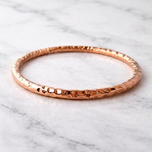 May include: A hammered copper bangle bracelet. The bracelet has a textured surface and a circular shape. The copper has a warm, reddish-brown colour. The bangle is displayed on a white and grey marble surface.