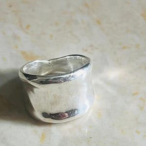 Puede incluir: Un anillo ancho de plata pulida con un perfil de borde irregular, orgánico y ondulado. La banda presenta una superficie lisa y reflectante con una forma ligeramente desigual y hecha a mano, creando una silueta única y no uniforme.
