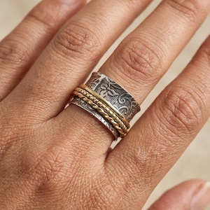 May include: A silver and gold spinner ring with a floral pattern. The ring features a wide, engraved silver band with a detailed floral design, and several thinner gold bands that spin freely around the silver band.