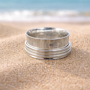 May include: A silver ring with a textured band and multiple thin, grooved bands. The ring is set on a sandy beach with the ocean in the background. The ring's design is simple and elegant, suitable for everyday wear.