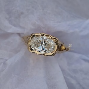 May include: A gold ring featuring two oval-cut diamonds set side by side. The diamonds are clear and sparkling, set in a textured gold band. The ring is displayed on a white, textured surface.