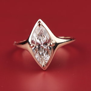 Könnte beinhalten: Ein goldener Ring mit einem Diamanten im Marquiseschliff, der in einer einzigartigen, geometrischen Lünette gefasst ist. Der Ring hat eine polierte Oberfläche und kleine goldene Akzente um den Diamanten. Der Hintergrund ist einfarbig rot.