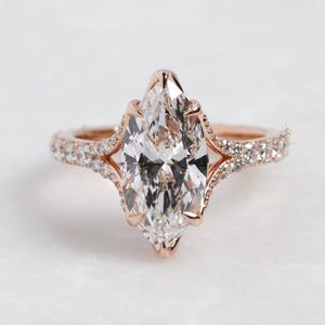 May include: Rose gold engagement ring featuring a large, marquise-cut diamond as the centerpiece. The band is adorned with smaller, pavé-set diamonds, adding sparkle. The ring's design includes intricate, openwork details on the sides, enhancing its elegant appearance.