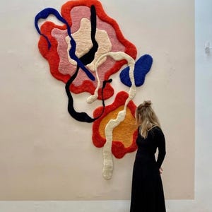 May include: Abstract wall art featuring a vibrant, multi-colored design. The artwork showcases a layered composition of red, pink, orange, blue, black, and white shapes. The textured piece is mounted on a light-colored wall, creating a striking visual contrast.