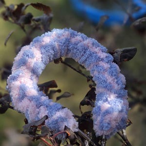 May include: A plush, circular wristlet keychain crafted from soft, fuzzy yarn in a variegated pattern of light blue and pale pink. The accessory features a silver-toned metal lobster clasp attached to one end for secure fastening to keys or bags.