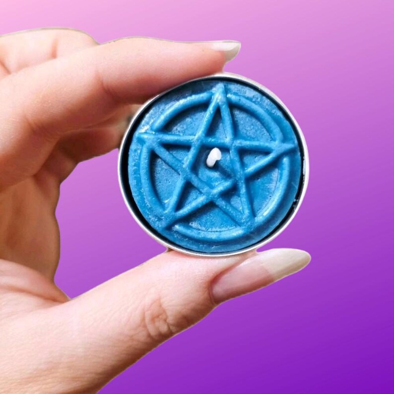 May include: A round blue wax candle featuring a raised pentagram design on its surface. The candle is housed in a thin metal container and has a single white wick positioned in the center of the star, creating a distinct geometric pattern.