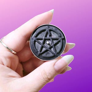 May include: A round black tea light candle featuring a raised pentagram design on its surface. The candle is contained within a thin silver-colored metal casing and has a small white wick centered in the middle of the star symbol.