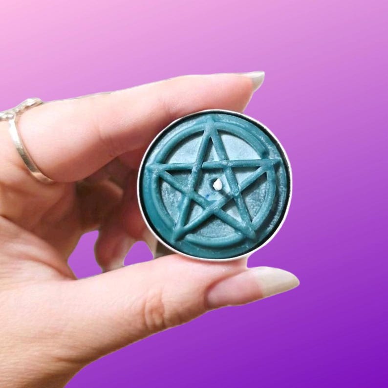 May include: A round teal wax melt featuring a raised pentagram design on its surface. The wax is contained within a thin silver-colored metal casing, and a small central wick hole is visible in the middle of the star symbol.