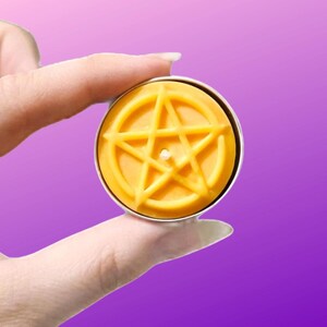 May include: A round yellow wax candle in a metal tin, featuring a raised pentagram design on the surface. The candle has a small, centered wick and is held between fingers against a solid purple background, showcasing its circular shape.