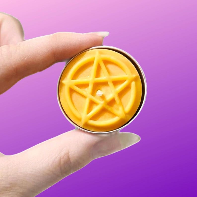 May include: A round yellow wax candle in a metal tin, featuring a raised pentagram design on the surface. The candle has a small, centered wick and is held between fingers against a solid purple background, showcasing its circular shape.