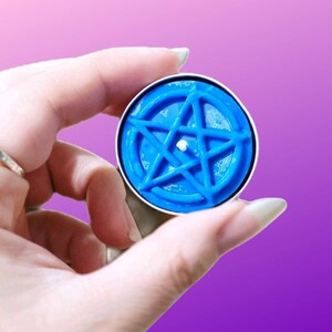 May include: A circular blue wax candle featuring a raised five-pointed star pentagram design on its surface. The candle is housed in a simple metal container and has a small white wick centered within the star shape, providing a clear view.