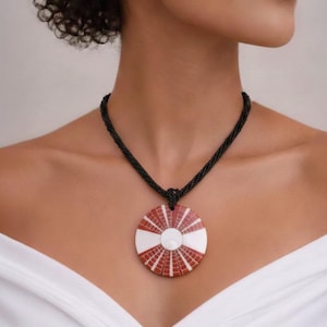 Può includere: Una collana con un pendente rotondo con un motivo geometrico rosso e bianco, sospeso a un cordino nero. La collana è indossata intorno al collo.